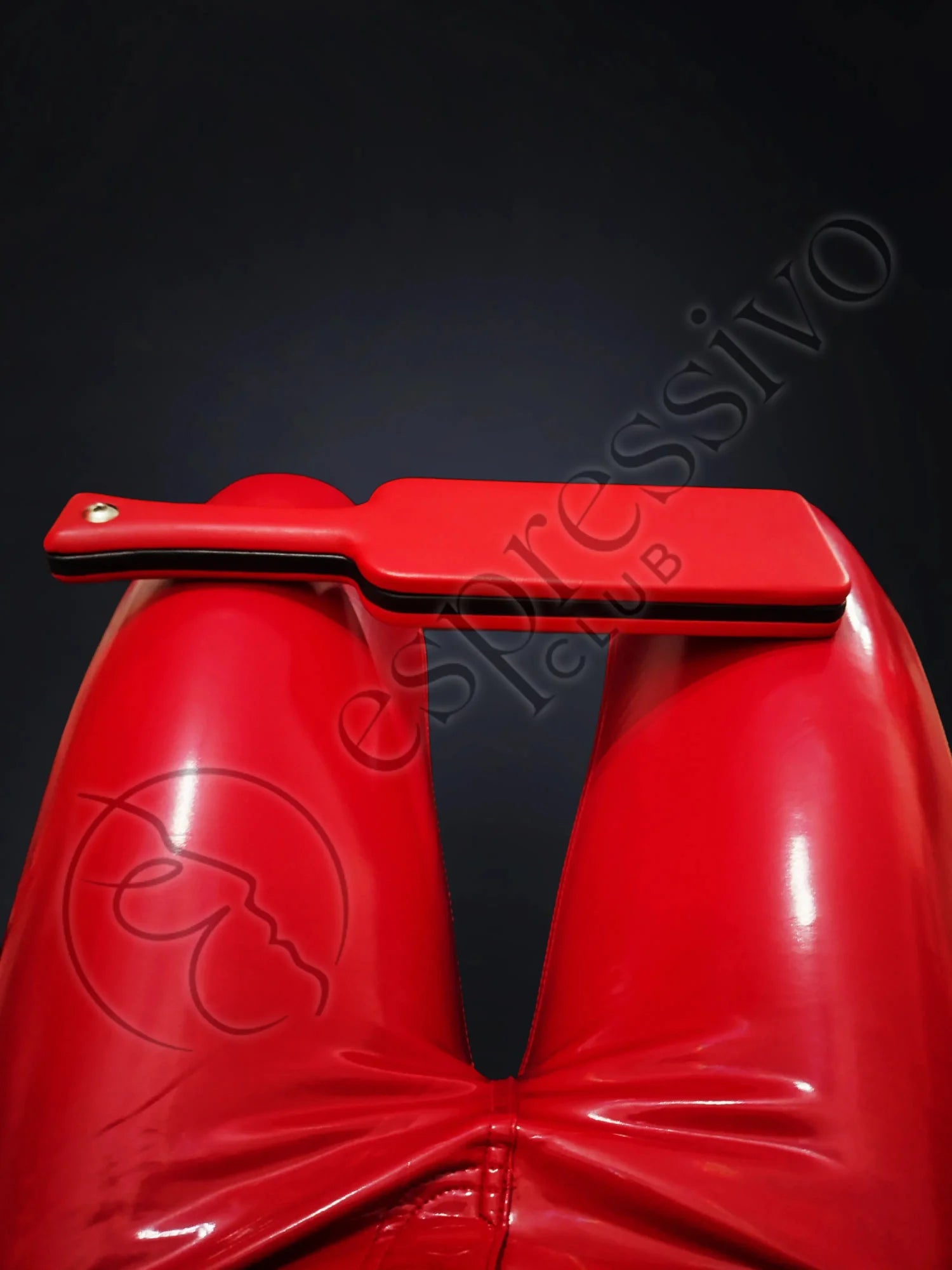 Red BDSM leather covered three layered wooden spanking paddle on the knees of fetish model in RED vinyl leggings.