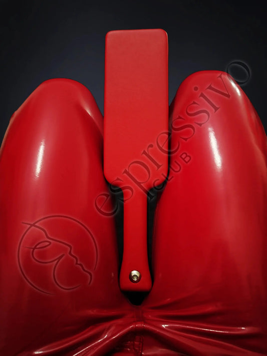 Red BDSM leather covered three layered wooden spanking paddle between the legs of fetish model in RED vinyl leggings.