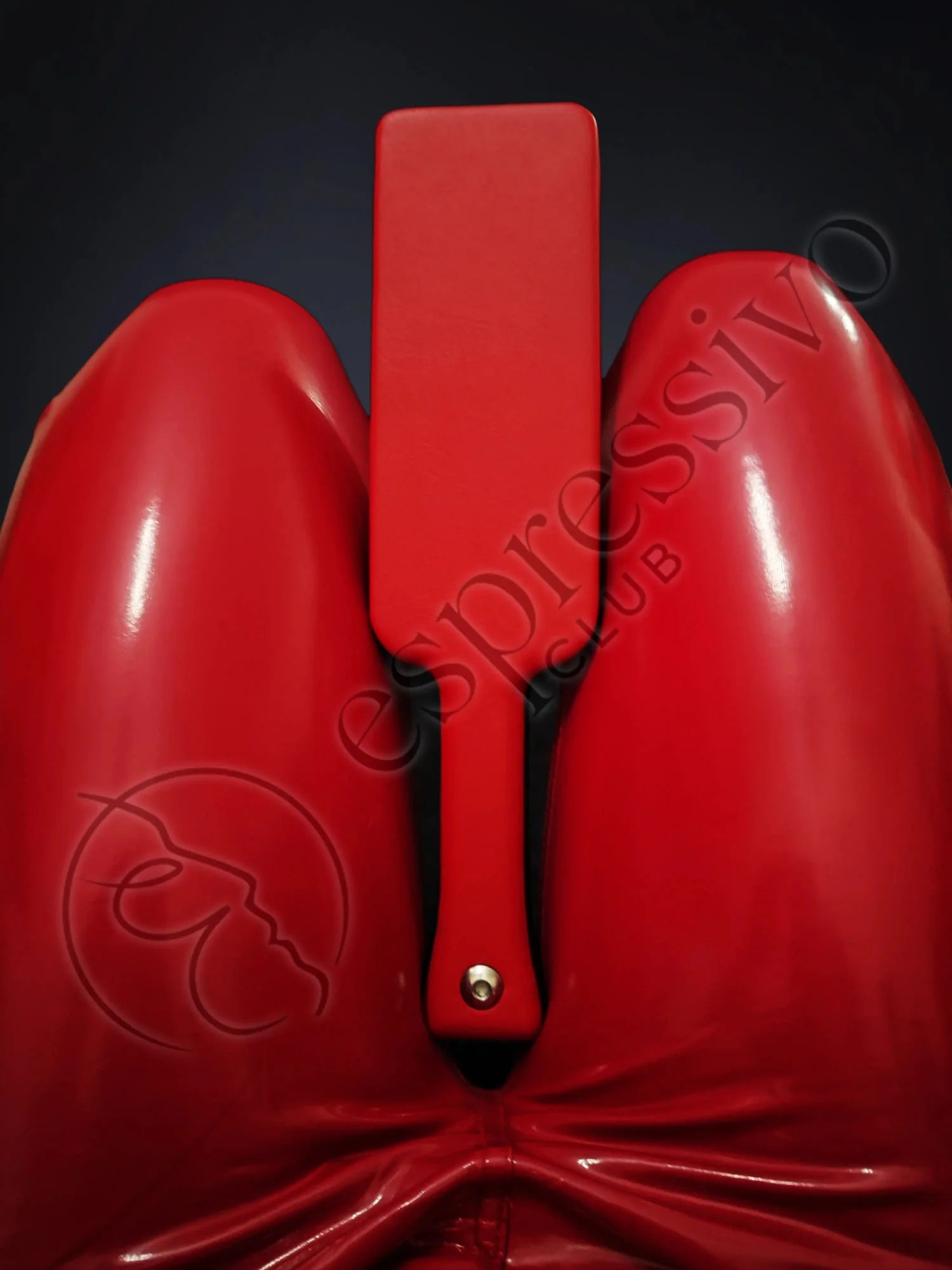 Red BDSM leather covered three layered wooden spanking paddle between the legs of fetish model in RED vinyl leggings.
