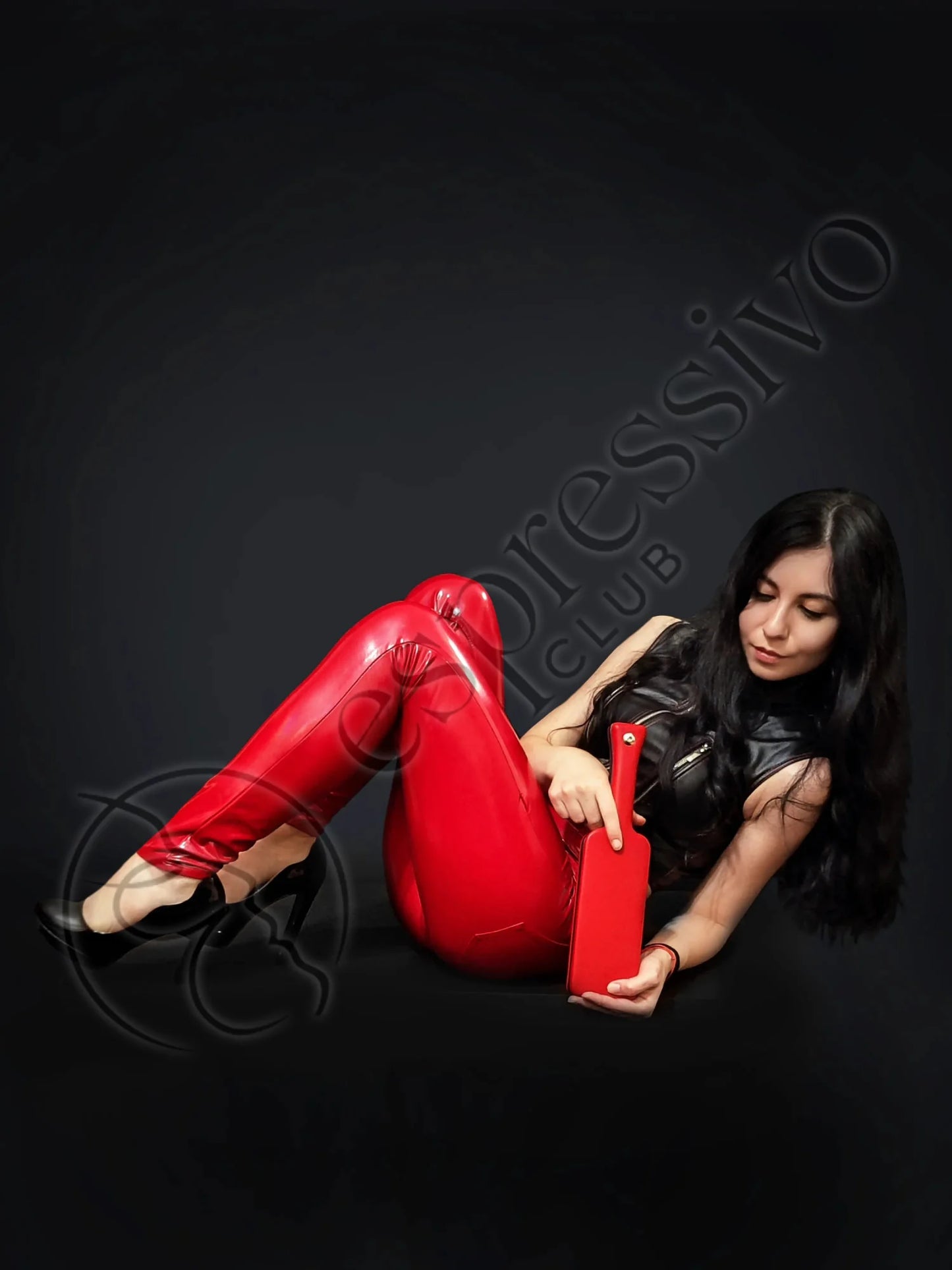 Fetish model in black leather top and red vinyl leggings holding a Red BDSM leather covered three layered wooden spanking paddle.